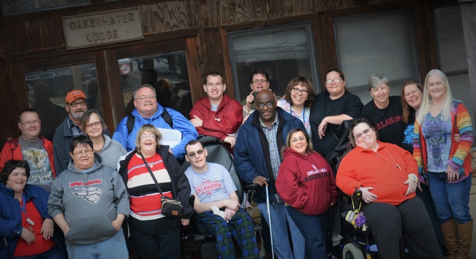 A group of men and women with disabilities stand outside in front of a wooden lodge.